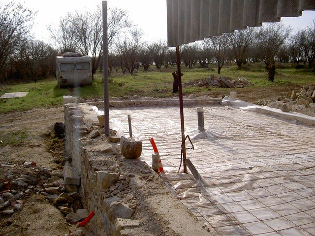 Dalle de béton fraîche sur treillis d'armature et film, bordée d'un muret de pierre ; caravane et arbres nus à l'arrière-plan.