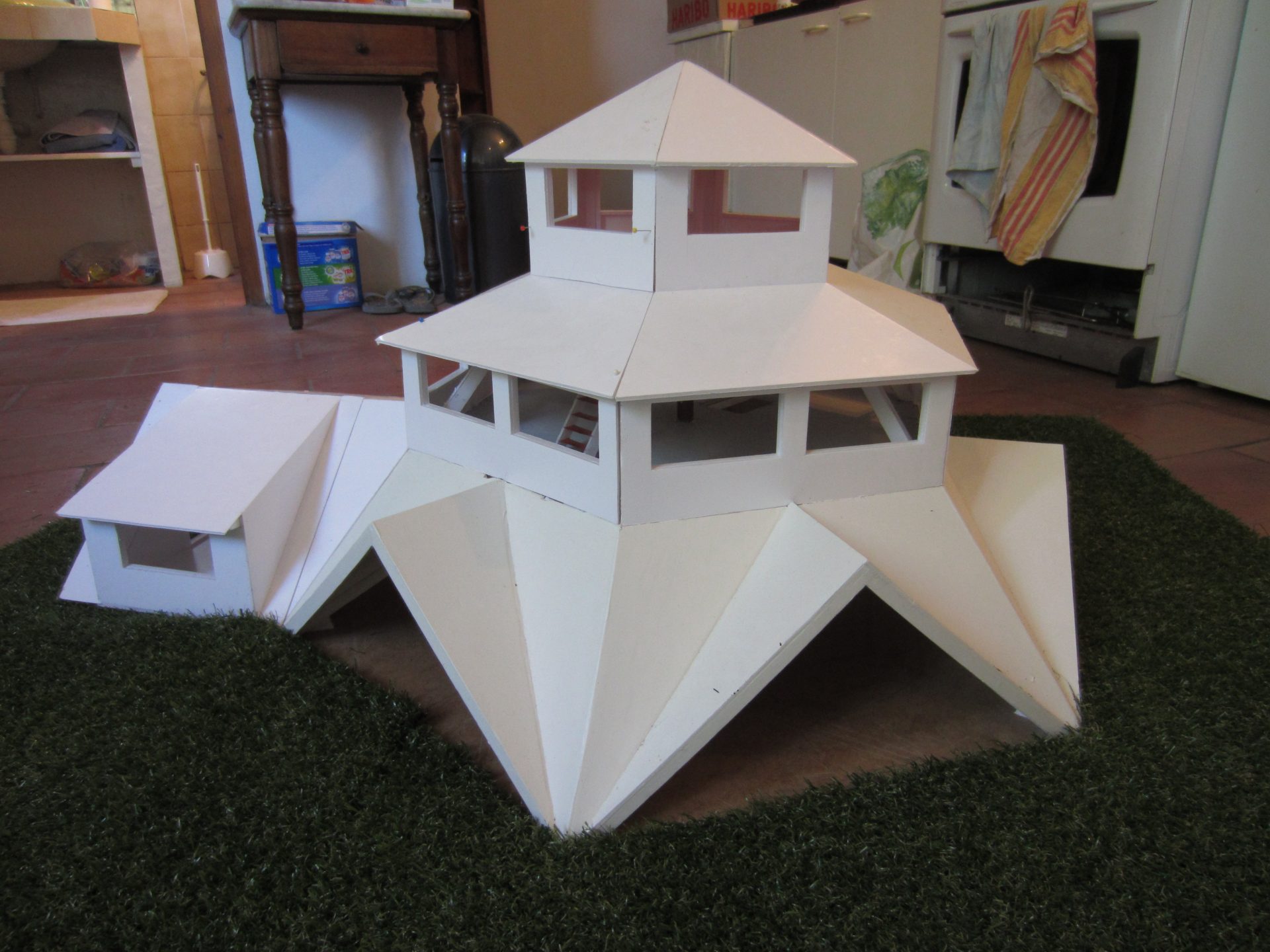 Maquette en carton blanc d'un pavillon à toiture facettée en gradins, posée sur un tapis d'herbe synthétique, en intérieur.